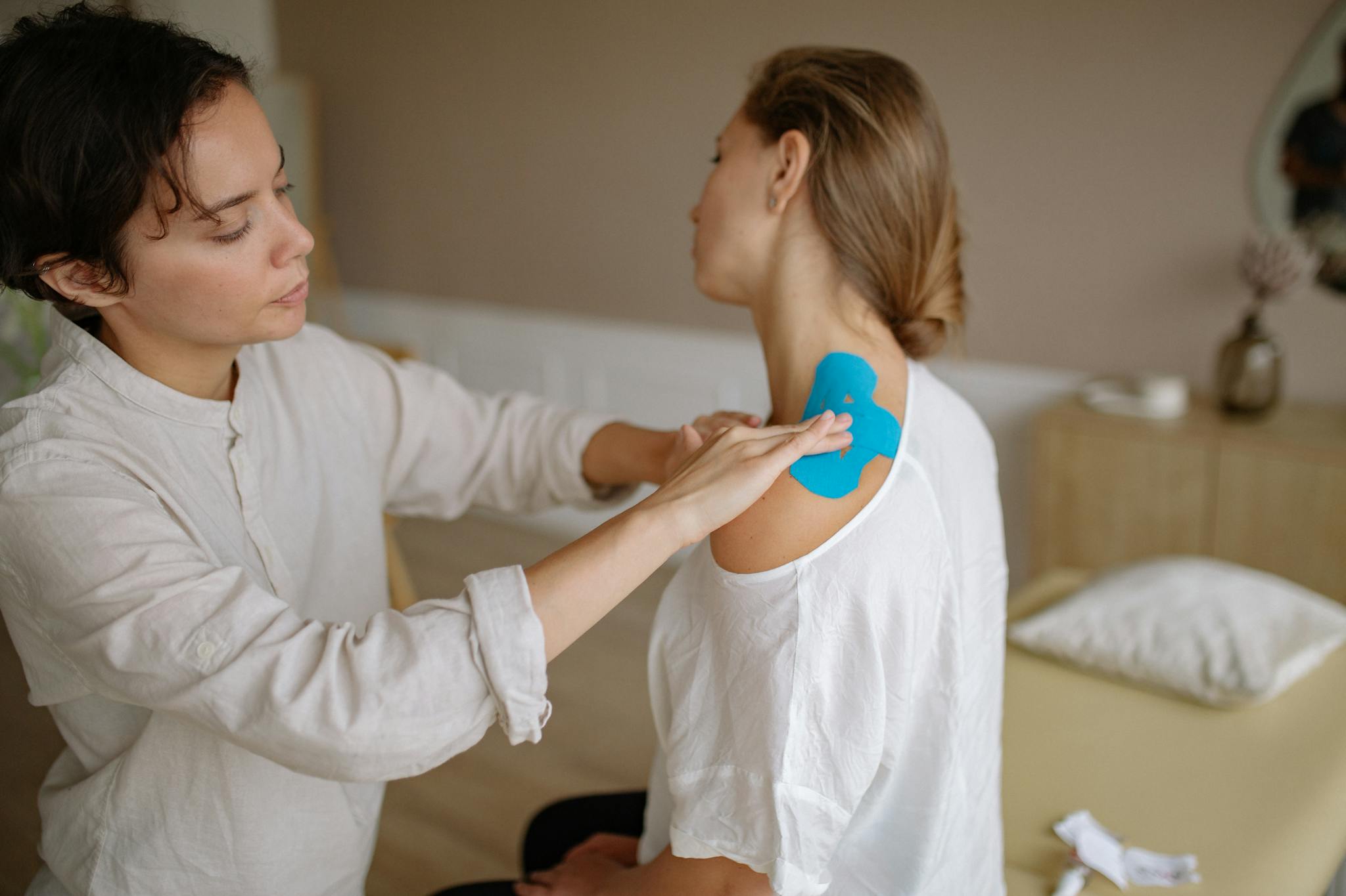 A physiotherapist applies blue kinesiology tape to a patient's shoulder for muscle support.